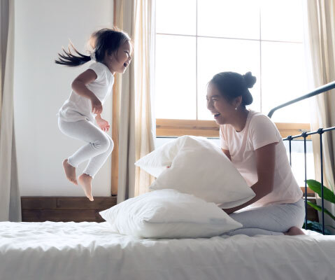 child jumping on bed as woman sits on the bed and laughs while holding pillows